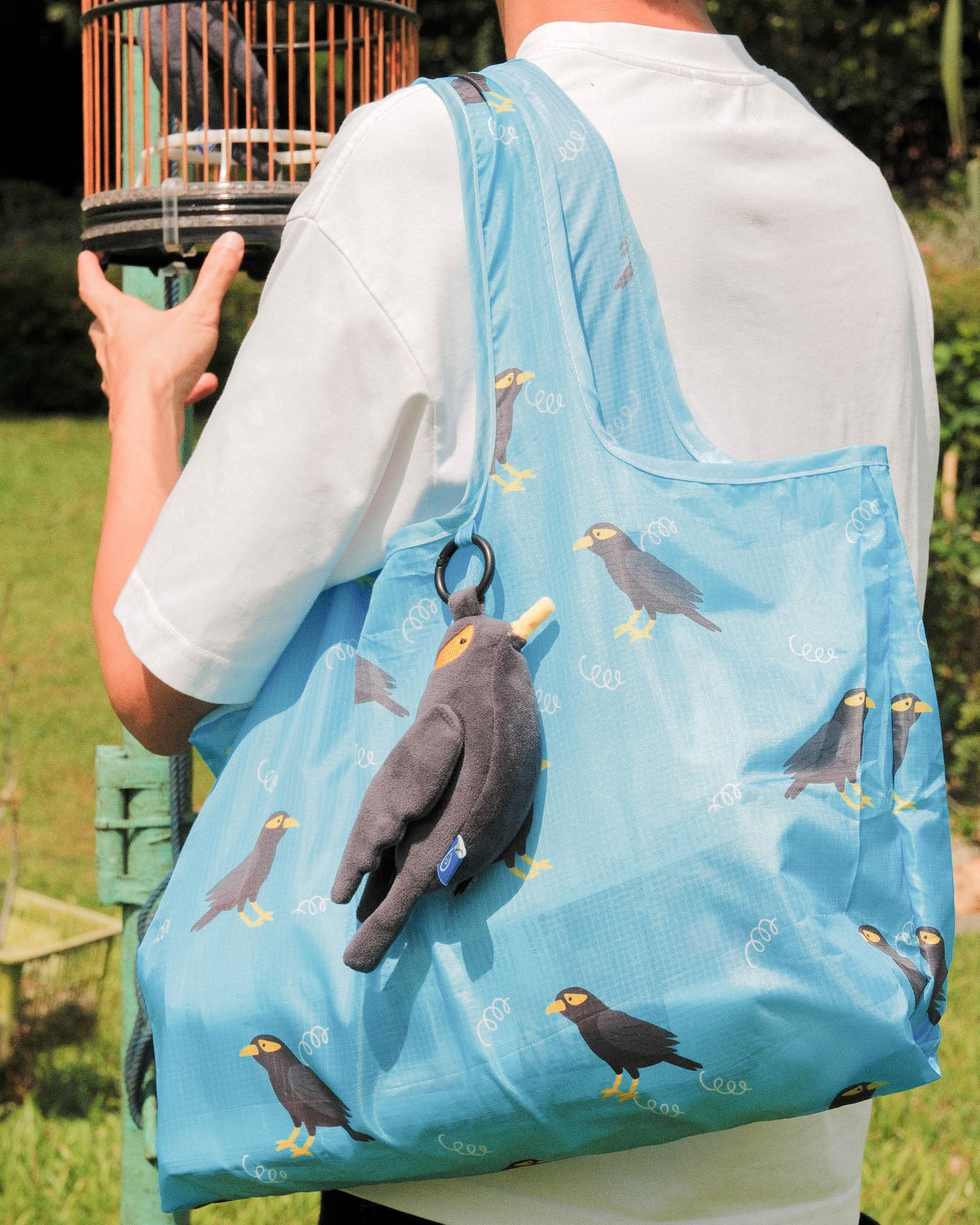 Person carrying a mynah bird plushie with blue reusable bag with bird design outdoors