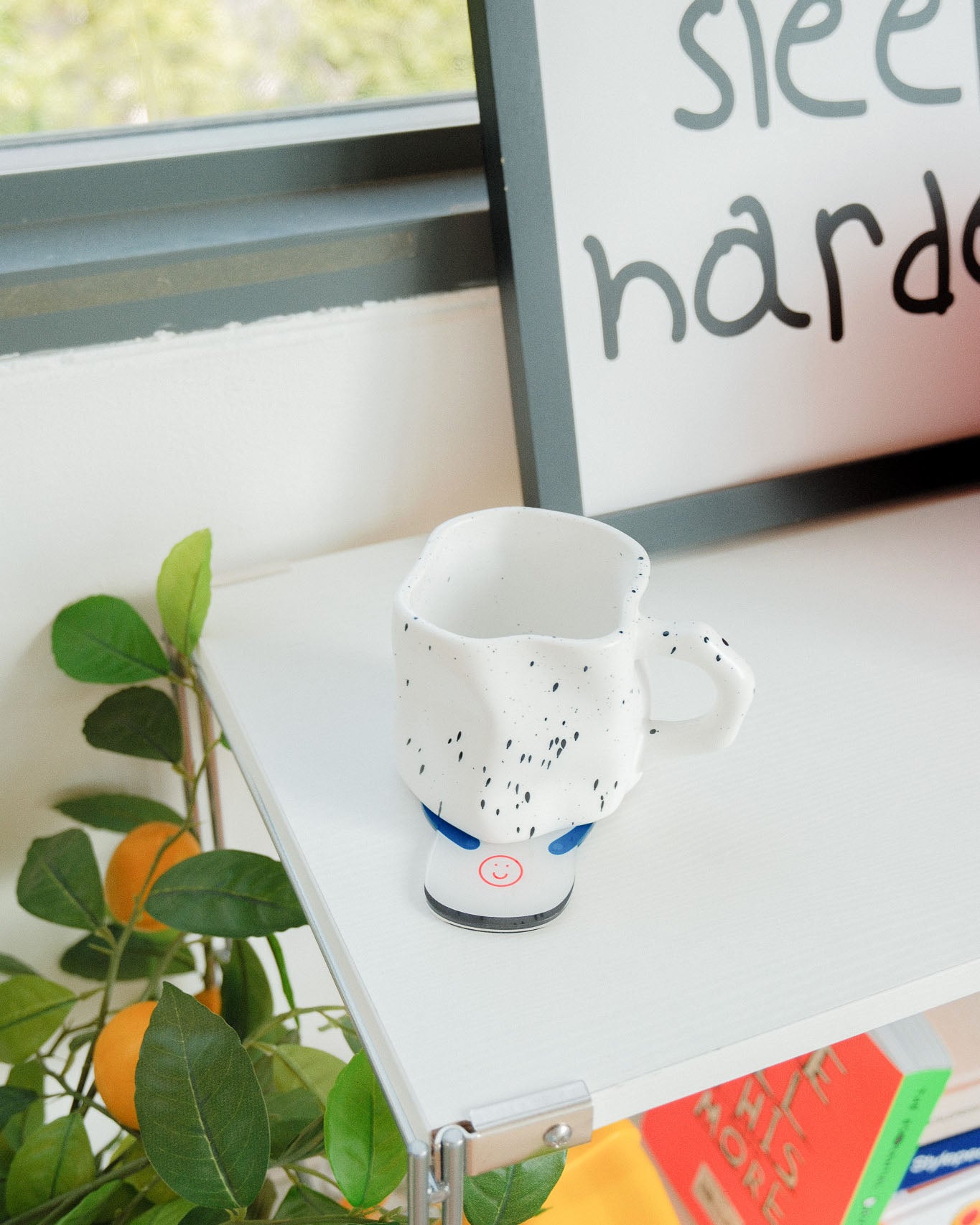 White mug with black speckles on a windowsill with plants and a framed picture in the background.